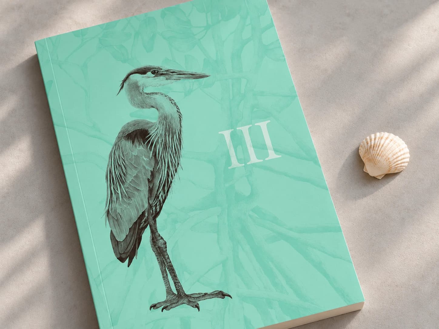 Close-up of the tilted aqua Breakwater Book III with heron and mangrove artwork, beside a small seashell